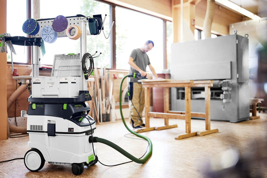Festool CLEANTEC CTM 36 EI Absaugmobil in Werkstatt mit Zubehöhr und Schlauch, Handwerker mit Elektrowerkzeug im Hintergrund